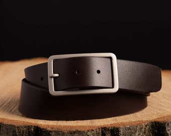 May include: A brown leather belt with a silver buckle. The belt is folded in half and sitting on a wooden surface.