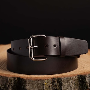 May include: A brown leather belt with a silver buckle. The belt is folded in half and resting on a piece of wood.