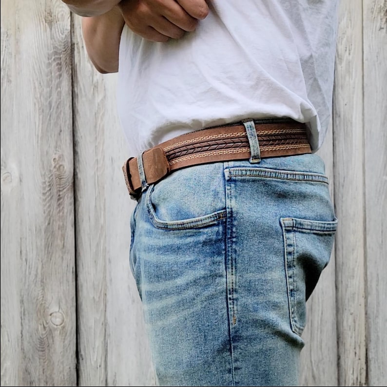 May include: A brown leather belt with a woven design and a rectangular buckle. The belt is worn with blue jeans and a white t-shirt. The belt is approximately 1.5 inches wide.