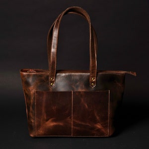 May include: A brown leather tote bag with two top handles and a zip closure. The bag has a distressed look and a front pocket.