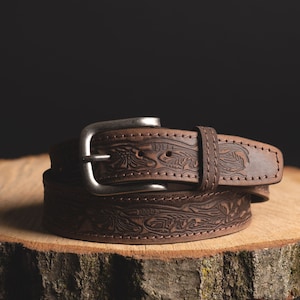 May include: A brown leather belt with an intricate floral design and a silver buckle. The belt is on a wooden surface.
