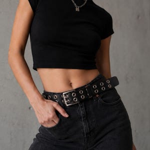 May include: Black leather belt with silver-tone grommets and a silver buckle. The belt is worn with black denim jeans and a black crop top. A silver necklace with a small lock pendant is also visible.