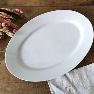 Op de afbeelding: Een witte ovale serveerschaal op een houten oppervlak. De schaal is gemaakt van keramiek en heeft een gladde, glanzende afwerking.