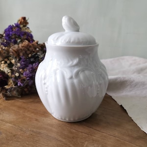 Könnte beinhalten: Weiße Porzellan-Zuckerdose mit Deckel und aufwendigem Blumendesign. Die Schale hat eine abgerundete Form und eine glatte, glänzende Oberfläche.