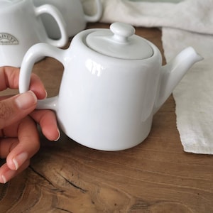Puede incluir: Tetera de cerámica blanca con tapa y asa. La tetera está sobre una superficie de madera.