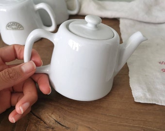 May include: White ceramic teapot with a lid and a handle. The teapot is sitting on a wooden surface.
