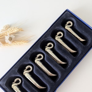 May include: A set of six silver-toned swan-shaped decorative items, presented in a navy blue box with individual compartments. The swans are arranged in a row, and a small bundle of dried wheat and flowers is in the background.