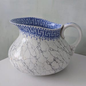 May include: A white and blue ceramic pitcher with a marble pattern and a blue Greek key design around the rim. The pitcher has a rounded body and a curved handle.