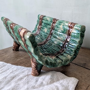 May include: A decorative ceramic bowl with a distinctive design. The bowl is green with brown detailing, reminiscent of a woven basket. It has a curved shape and is supported by three small legs. The bowl is placed on a wooden surface.