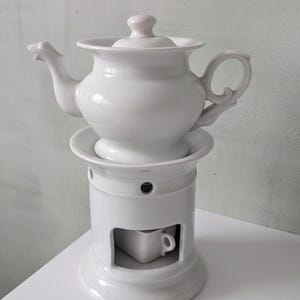 Op de afbeelding: Een witte keramische theepot met bijpassende deksel en ingebouwde warmer. De theepot heeft een gebogen tuit en handvat. De warmer heeft een cilindrische basis met een kleine opening voor een waxinelichtje.