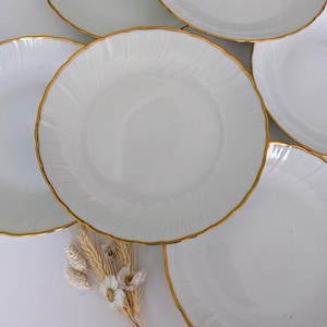 May include: A set of white ceramic plates with a gold trim. The plates feature a wavy edge design and are arranged in a circular pattern. A small bunch of dried flowers is placed near the plates.