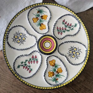 May include: A round ceramic oyster plate with a cream-colored base and a yellow and black patterned rim. The plate features six recessed, shell-shaped sections decorated with hand-painted floral motifs and a central circular well with pink rings.