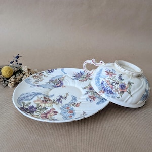 May include: A vintage teacup and saucer set. The white porcelain cup and saucer feature a floral design in blue, purple, and pink. The cup has a curved handle, and the saucer has a unique shape with a handle.