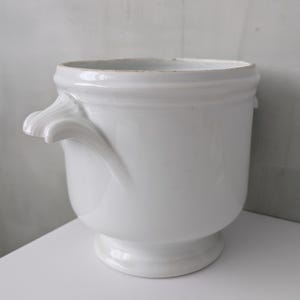 Puede incluir: Una maceta de cerámica blanca con base de pedestal y dos asas decorativas. La maceta tiene una abertura ancha y un acabado liso y brillante. La forma general es un cilindro redondeado, ligeramente cónico.