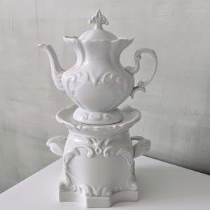 Può includere: Una teiera in ceramica bianca con coperchio abbinato, posta su uno scaldavivande decorativo. La teiera presenta dettagli floreali e a volute ornati, con manico e beccuccio. Lo scaldavivande ha un design simile, con manici su entrambi i lati.