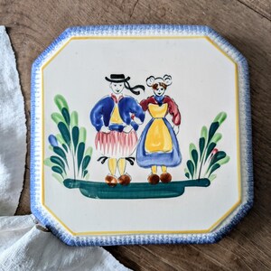 Può includere: Piastrella in ceramica con bordo blu e giallo raffigurante due figure in abiti tradizionali. Le figure sono su una base verde con fogliame verde stilizzato. La piastrella ha una forma ottagonale.