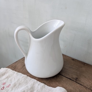 Puede incluir: Una jarra de cerámica blanca con un asa curva y un pico vertedor. La jarra está sobre una superficie de madera junto a un paño blanco con texto rojo. El fondo es una pared azul claro.