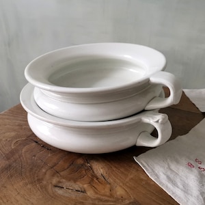 Peut inclure: Deux pots de chambre en céramique blanche empilés l'un sur l'autre. Les pots de chambre ont une forme arrondie et une poignée de chaque côté.