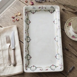 Puede incluir: Una bandeja rectangular de cerámica blanca con borde de guirnalda floral, una taza de té con plato a juego, una servilleta de lino beige, un tenedor, un cuchillo y dos pinzas para azúcar de plata junto a pétalos de rosa y lavanda seca.
