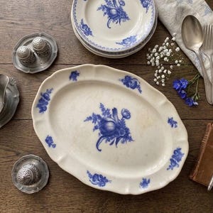 Könnte beinhalten: Ein Set aus cremefarbenem Keramikgeschirr mit blauen Blumenmustern, bestehend aus einer ovalen Platte, kleinen Tellern, einer Sauciere und Salz- und Pfefferstreuern aus Metall, ergänzt durch einen Löffel, eine Gabel, eine Serviette und einen Füllfederhalter.