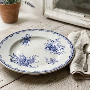 Peut inclure: Une assiette ronde en céramique à fond blanc ornée de motifs bleus de type transfert. Le bord présente un motif de feuilles en continu, tandis que le centre est décoré de compositions florales bleues et d'une illustration d'oiseau.
