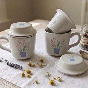 Op de afbeelding: Twee off-white keramische theemokken met bijpassende deksels en uitneembare theezeefjes. Elke mok is voorzien van een beschilderd ontwerp van een blauwe pot met roze bloemen en groene bladeren. Inclusief een glazen potje met gedroogde kruiden en een zilveren lepel.