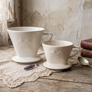 Może przedstawiać: Dwa białe ceramiczne zaparzacze do kawy z uchwytami. Większy zaparzacz ma wytłoczony napis Melitta 123, a mniejszy napis Melitta 101. Oba posiadają szeroką, okrągłą podstawę oraz żebrowaną powierzchnię wewnętrzną ułatwiającą przepływ kawy.