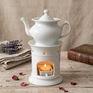 Op de afbeelding: Een witte keramische theepot staat op een bijpassende witte keramische theelichthouder. De cilindrische voet heeft een uitsparing met daarin een klein brandend waxinelichtje. De theepot heeft een deksel, een gebogen handvat en een tuit.