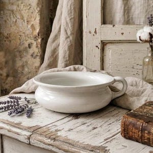 May include: A white ceramic chamber pot with a single side handle and a wide, rounded basin. The vessel features a smooth, glossy finish and a low-profile design, positioned alongside a sprig of dried purple lavender on a weathered surface.