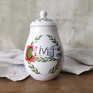 Könnte beinhalten: Weiße Keramik-Oliven-Dose mit grünem Blattdesign und dem Wort "OLIVES" in schwarzer Schrift. Die Dose hat einen Deckel mit grünem Blattdesign.