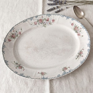 Könnte beinhalten: Eine ovale Servierplatte aus Keramik mit cremefarbener Basis und einer feinen Craquelé-Glasur. Der Rand ist mit einer blauen geometrischen Bordüre sowie mit rosa und roten Rosen samt grünen Blättern verziert.