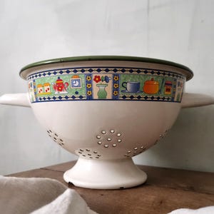 Op de afbeelding: Witte emaille zeef met een groene rand en een kleurrijk ontwerp met een keukenthema. De zeef heeft twee handvatten en een bodem met gaten.