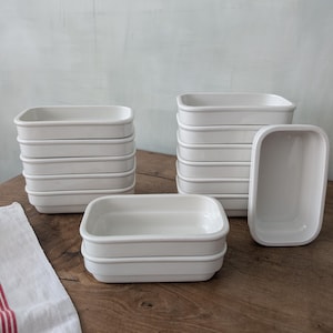 Op de afbeelding: Verschillende stapels witte rechthoekige keramische schalen met afgeronde hoeken zijn gerangschikt op een houten oppervlak. De schalen hebben een eenvoudig, strak ontwerp met een licht verhoogde rand. Een witte doek met rode strepen is zichtbaar.