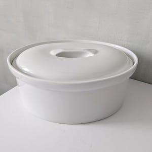 May include: A white ceramic casserole dish with a lid. The dish is round with a slightly flared base and a matching lid with a handle. The dish is sitting on a white surface.