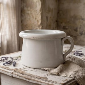May include: A white enamel chamber pot with a wide, rounded rim and a single side handle. The surface of the vessel shows signs of wear, including minor chipping along the top edge, revealing the underlying metal base of the object.