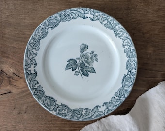May include: White ceramic plate with a blue floral design. The plate features a central rose motif and a decorative border of scrolling leaves and flourishes. The plate is round and appears to be vintage.