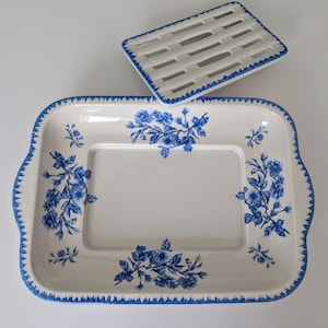 May include: A white rectangular serving dish with blue floral designs and a matching trivet. The dish has a scalloped edge with blue trim. The trivet has a woven pattern and blue trim. Both items are made of ceramic.