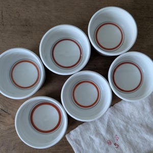 以下が含まれることがあります： 赤い輪郭と黒い輪郭のデザインが施された白い陶器のボウルが6つ。ボウルは木製の表面に円形に配置されています。