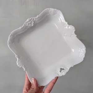 Puede incluir: Una fuente blanca de forma cuadrada con bordes ornamentados y enrollados. La fuente tiene un acabado brillante y detalles calados decorativos. El diseño recuerda a la vajilla vintage. La fuente está sostenida por una mano.