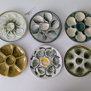 May include: Six round ceramic oyster plates arranged in two rows. The plates feature various glaze colors including white, green, yellow, and brown, with molded shell-shaped indentations for serving oysters and a central well for condiments.