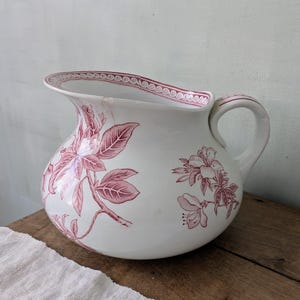 May include: White ceramic jug with a rounded body and a handle. The jug is decorated with a floral pattern in shades of red. The rim features a decorative border. A classic piece for serving beverages or as a decorative item.