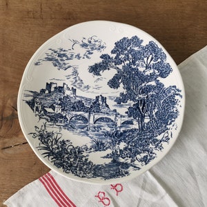 Könnte beinhalten: Weißer Keramikteller mit blauem Landschaftsdesign mit Brücke, Bäumen und Burg. Der Teller hat einen dekorativen Rand und liegt auf einer Holzoberfläche mit einem weißen Tuch.