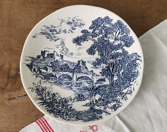 May include: White ceramic plate with a blue landscape design featuring a bridge, trees, and a castle. The plate has a decorative rim and is set on a wooden surface with a white cloth.