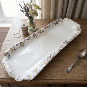 Puede incluir: Una bandeja de servir rectangular y alargada de cerámica blanca con un delicado patrón floral en tonos morados, amarillos y verdes a lo largo de los bordes festoneados. La bandeja incluye detalles de asas ornamentadas en ambos extremos.