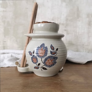 Könnte beinhalten: Eine weiße Keramikdose mit einem Blumendesign in Blau, Braun und Orange. Die Dose hat einen Korkdeckel und einen Holzlöffelhalter an der Seite.