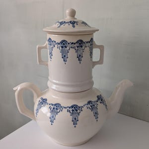 Puede incluir: Cafetera de cerámica color crema con un patrón decorativo azul. La cafetera tiene una base bulbosa, un pico y un asa. Una pieza cilíndrica separada con asas y una tapa se encuentra encima. El patrón azul se repite.