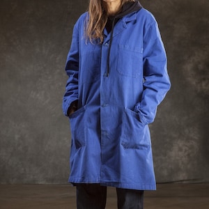 May include: A blue work jacket with a hood and two large pockets. The jacket is worn over a pair of dark blue jeans and black sneakers.