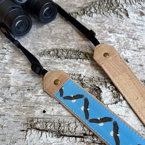 May include: A camera or binocular strap featuring a light blue fabric center with a repeating pattern of bald eagles in flight. The strap ends are made of tan cork material, secured with brass-colored metal rivets and black adjustable nylon webbing.