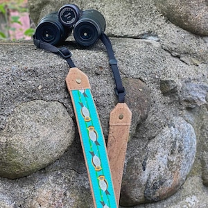 Może przedstawiać: Para czarnych lornetek z brązowym i niebieskim paskiem na aparat. Lornetki mają nadrukowany napis "Vortex 10x42 Talon".