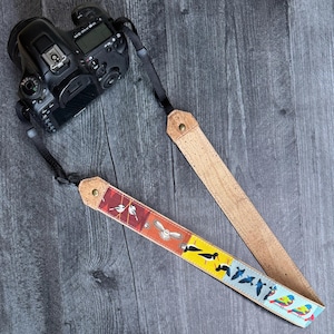 Puede incluir: Una cámara DSLR negra con una correa decorativa. La correa es de corcho y presenta un diseño de pájaros coloridos. La correa está conectada a la cámara con correas de nailon negro.